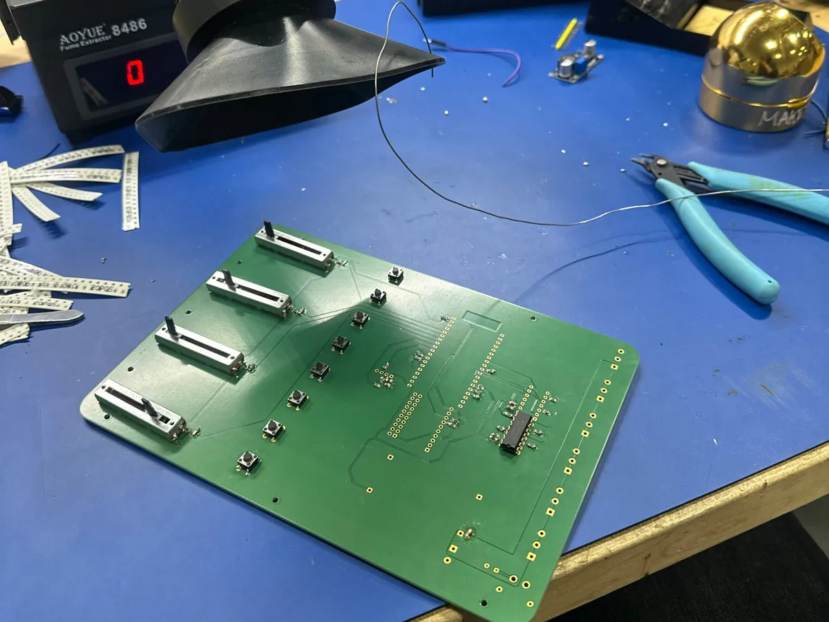 The custom PCB on a blue bench mat, partly assembled with four slide potentiometers and tactile buttons, next to a fume extractor, solder wire, flush cutters and strips of SMD parts